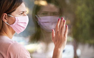 Wall Mural - Young woman in virus mask looking sad from the window behind glass pane, touching it with hand. Quarantine or stay at home to be safe during coronavirus covid-19 outbreak concept