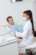 © LIGHTFIELD STUDIOS - african american dentist in medical mask and man looking at face shield in clinic
