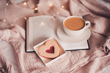 Fresh Cup Of Coffee With Heart Shape Cake On Open Book In Bed Over Lights Closeup. Good Morning. Breakfast. Valentines Day.
