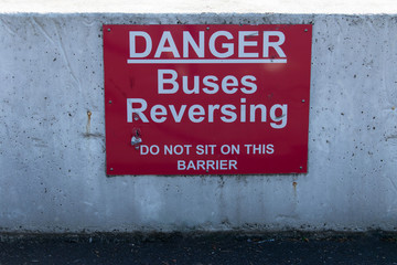 A bold red and white Danger warning sign stating - Buses Reversing; Do Not sit on this barrier