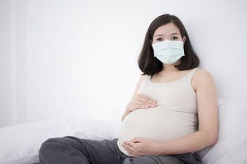 Pregnant asian woman in medical face mask to protect Covid-19 (Coronavirus) and PM 2.5 sitting on bed and touching her big belly 