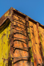 A Old Rusty Ladder Leading To The Top Of A Weathered And Rusty Yellow, Red And Orange Construction Container.  The Close Up Image Is Looking Upwards And The Background Is A Cloudless Bright Blue Sky.