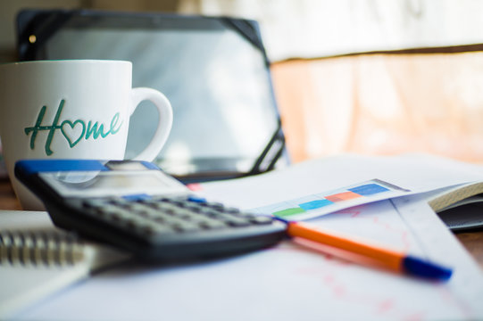 On the home table are stock and finance charts with a calculator, a coffee mug, a pencil, a marker pen, two notebooks, a smartphone and a tablet for remote work. Work at home.