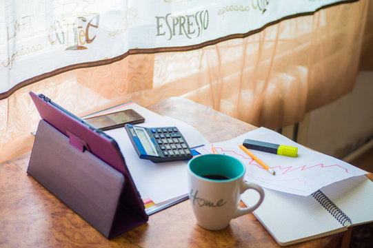On the home table are stock and finance charts with a calculator, a coffee mug, a pencil, a marker pen, two notebooks, a smartphone and a tablet for remote work. Work at home.