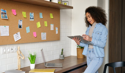 Wall Mural - Smiling africanteen girl college student studying from home checking homework in notebook, elearning, stand at home office desk, making notes. Young black woman remote learning remote school lessons.