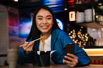 Photo of asian woman eating chinese noodles and using cellphone in cafe