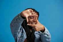 Happy Black Female With Braids In Denim Jacket Smiling And Looking At Camera While Framing Face With Hands Against Blue Background