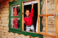 Cheerful Black Child In Red Shirt And White Pants Looking At Camera Through Open Window Of Rural Wooden House