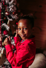 Side View Of Cheerful Little Black Girl In Red Shirt Looking At Camera And Smiling While Standing Near Decorated Christmas Tree At Home