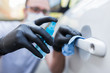 Man wearing protective mask and gloves while using small microfiber cloth and disinfectant alcohol sprayer to eliminate dirt or bacteria from car door handle.