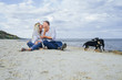 © Daria - Beautiful, attractive couple sitting on the sand by the sea, huging, kissing, petting dogs and laughing. In the foreground there are funny, shaggy dogs running, bitting each other and bothering.