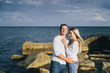 © Daria - Long shot. Attractive couple in bright clothes standing on stones in the sea against a blue cloudy sky. Lit by the soft evening sun. Looking forward, hug, kissing and enjoying beautiful evening