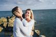 © Daria - Long shot. Attractive couple in bright clothes standing on stones in the sea against a blue cloudy sky. Lit by the soft evening sun. Looking forward, hug, kissing and enjoying beautiful evening
