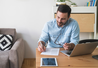 Wall Mural - Young businessman sitting at a table working at home on a laptop and writing on paper stock photo