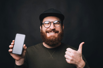 Close up portrait of a bearded man wearing glasses showing thumb up and his phone on black background.