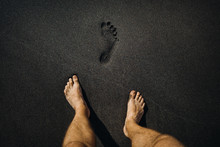 Close up of male footprints and feet walking on the volcanic black sand on the beach