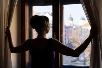 Woman back opening curtains looking from apartment room glass window with view of Lviv, Ukraine old market town square in elegant dress