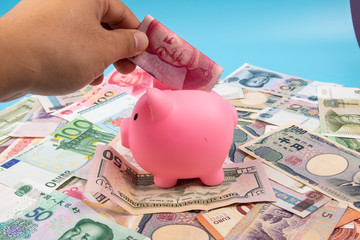 Man hand holding RMB YUAN bank note into piggy bank with World banknotes collection mixed on table, top view. Business and finance concept.