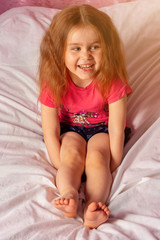 Wall Mural - Caucasian little girl with lush hair laughing while sitting on bed