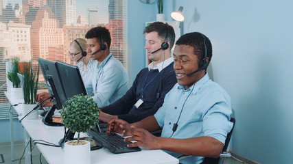 Wall Mural - Medium long shot of Black customer support agent working in busy call center by talking to the international client. His multiethnic team also taking calls.