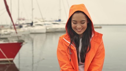 Wall Mural - Amazing asian smiling positive woman walking by harbor grant outdoors in orange coat.