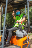 Fototapeta Panele - woman excavator on a small excavator with yellow warning vest wearing the sanitary protective mask that makes the gesture of everything ok