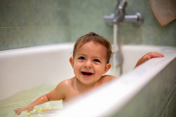 Wall Mural - Cute Caucasian baby taking a bath, smile and look up, resting on the side of the bath. water splashes, in the background a green bathroom in blur. close-up, soft focus