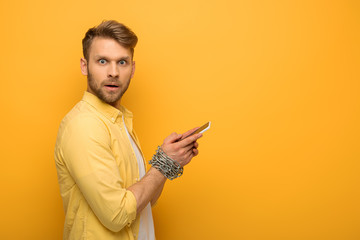 Side view of confused man with metal chain around hands holding smartphone and looking at camera on yellow background