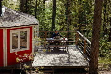 High angle view of middle aged man sitting at table on porch of country house surrounded by pine trees, drinking tea and working on laptop computer