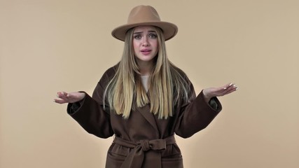 Poster - A puzzled confused woman wearing hat and coat is raising her hands isolated over the beige background in studio