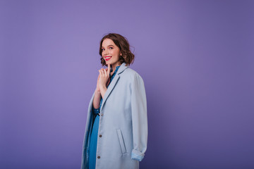 Wall Mural - Romantic white woman in elegant coat playfully posing on purple background. Indoor photo of joyful curly female model with short haircut.