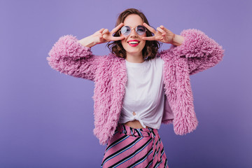 Joyful young lady in fluffy coat funny dancing on purple background. Indoor portrait of sensual white woman with wavy hair laughing to camera.