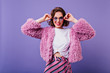 © Look! - Glamorous young woman with dark wavy hair touching her trendy glasses. Indoor portrait of pleasant caucasian girl in fluffy fur jacket isolated on purple background.
