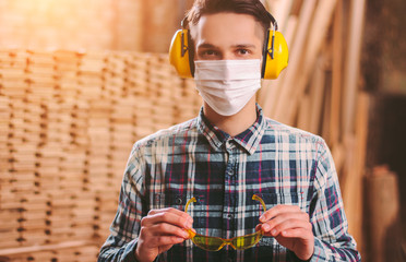 Wall Mural - Portrait of young carpenter in hearing protectors headphones and medical face mask wearing protective eyewear at workshop. Male craftsman at sawmill. Factory work at coronavirus COVID-19 quarantine