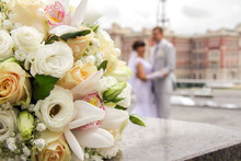 Bouquet Of Wedding Flowers In The Foreground. On The Background Of A Wedding Couple Are Standing In An Embrace. Photo Near The Registry Office On The Square