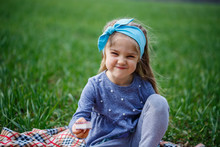 Little Girl Child Sits On The Bedspread And Eats Cookies And Marmalade, Green Grass In The Field, Sunny Spring Weather, Smile And Joy Of The Child, Blue Sky With Clouds