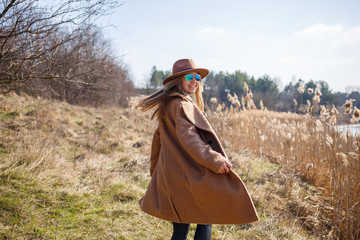 Wall Mural - A girl in a brown coat, hat and glasses walks in a park with a lake under the bright sun. Rejoices in life and smiles. The beginning of spring