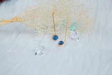 Earrings Made Of Blue Round Beads And Golden Yellow Metal.  Yellow Dry Grass Plants As A Background.  White Plush Cloth And Transparent Ice Ornaments.