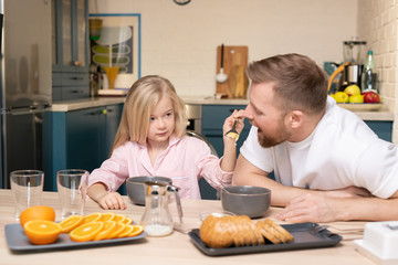 Wall Mural - Adorable little girl holding spoon with corn flakes by her father mouth by table