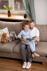 Wall Mural - Clever little girl in pajamas sititng on knees of her father while reading book