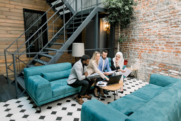 Group of young concentrated international business people discussing a new business project and important ideas, while sitting on blue couch and using laptop and digital tablet computers