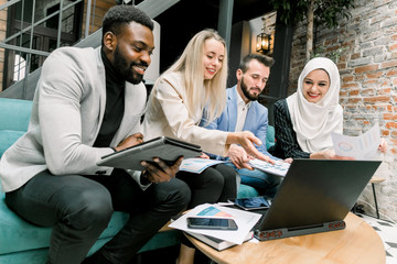 Side angle view of four young satisfied multiethnic business colleagues, smiling while looking together on the screen of laptop with good results of their joint financial project, sitting in office