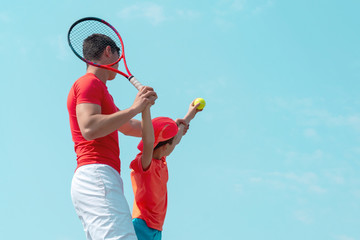 Wall Mural - A young tennis coach or instructor teaches a child a serving technique. Boy tennis player throws the ball up for serve. Kids sports school. Blue sky background. Banner. Copy space for text.