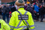 Fototapeta  - A male medical first responder walks along a street alone with people standing in the distance. The officer is wearing a bright yellow coat with grey reflective bands, black pants and a radio.