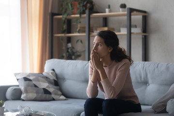 Thoughtful doubtful young woman sitting alone on couch, feeling nervous about hard decision. Stressed millennial girl thinking of loneliness psychological problem, regret about relationship mistake.