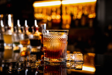 A cocktail in an old fashioned glass on a bar counter with a reflection, ice cubes around, bottles with bitters, bokeh lights, horizontal photo