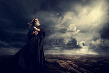 Monk Holding Bible Looking Up To God Sky Light, Old Priest In Black Robe In Storm Mountains