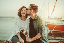 Loving Cheerful Couple Of Lovers Kiss While Sitting On A Bench Against The Background Of  Yachts Club At Sea