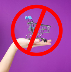 Wall Mural - Mini toy metal shopping trolley on female hand on purple background with prohibition sign.