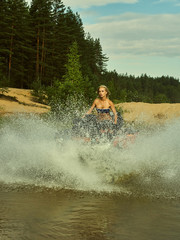 Wall Mural - Beautiful blonde girl on a ATV rides on water and sand mountains. Green forest all around. In summer season